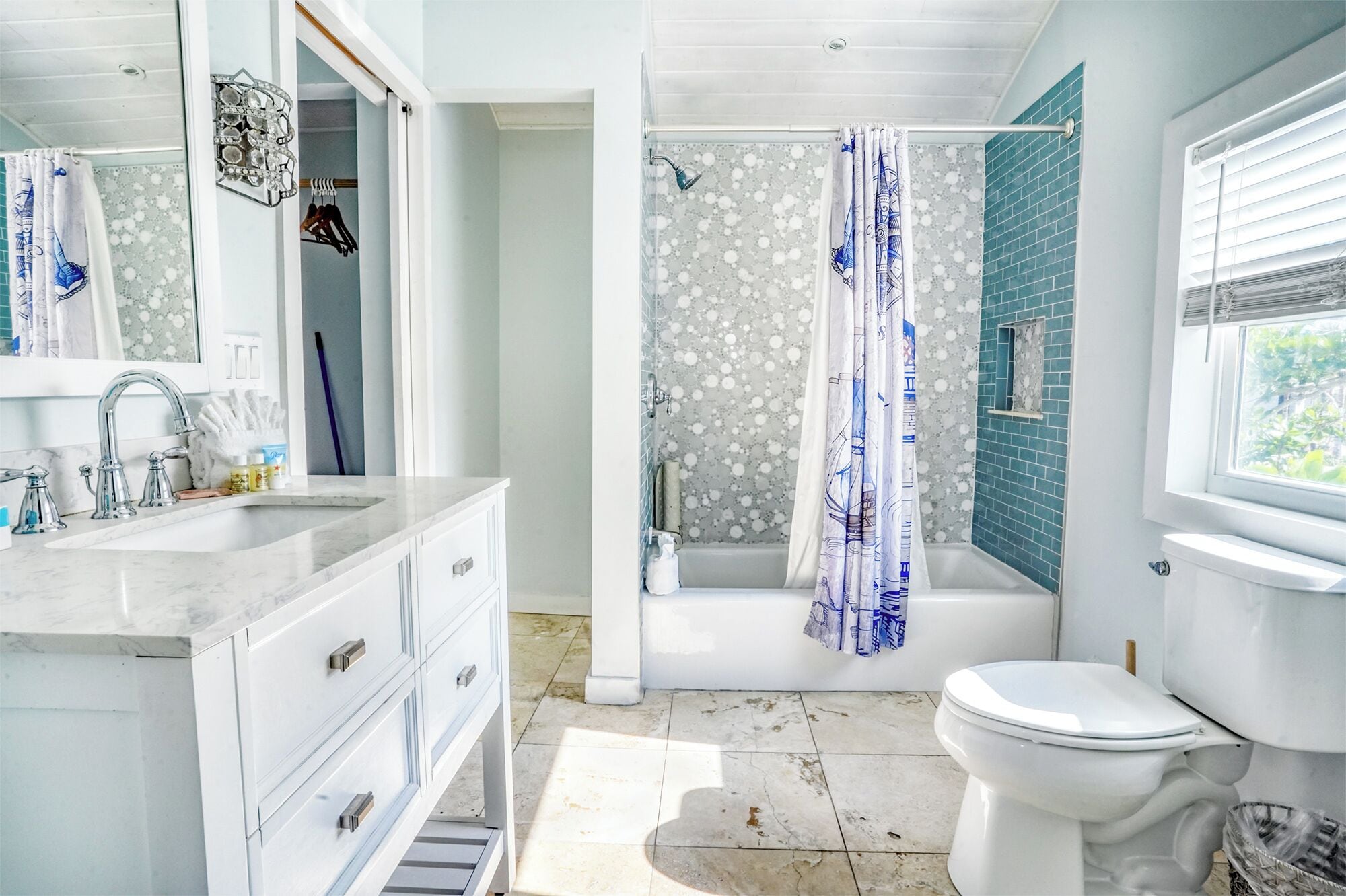 Une salle de bain avec un lavabo, un meuble-lavabo et des toilettes, tous de couleur blanche. On y trouve également une douche avec un rideau de douche bleu et une baignoire. Les murs sont recouverts de carreaux, et il y a une fenêtre avec des stores.