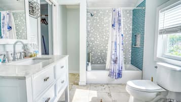 A bathroom with a white sink, vanity, and toilet. There is a shower area with a blue shower curtain and a bathtub. The walls are tiled, and there is a window with blinds.