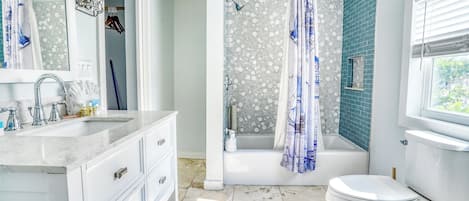 A bathroom with a white sink, vanity, and toilet. There is a shower area with a blue shower curtain and a bathtub. The walls are tiled, and there is a window with blinds.