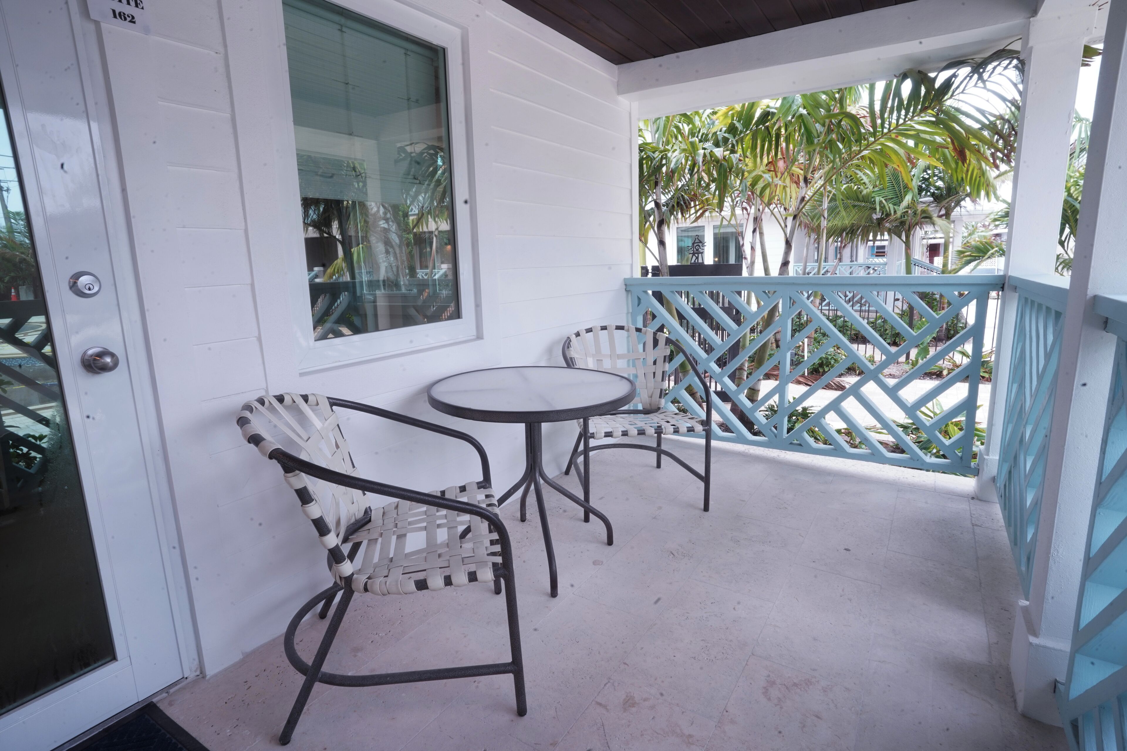 A covered outdoor area with a white door, two wicker chairs, a small round table, and a blue lattice railing.