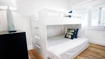 Una cama litera con colchón y almohadas blancos, una lámpara sobre una mesita de noche negra, y una ventana con persianas.