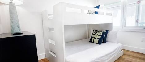 Una cama litera con colchón y almohadas blancos, una lámpara sobre una mesita de noche negra, y una ventana con persianas.