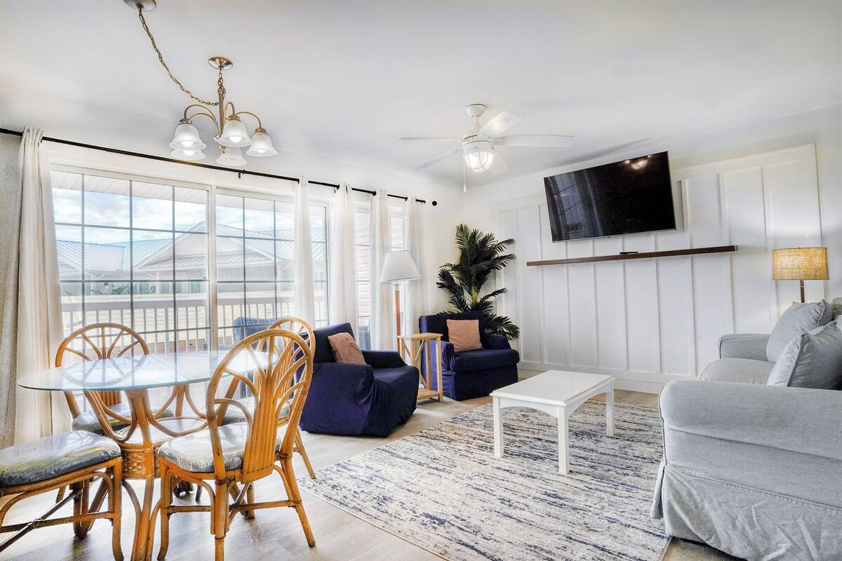 A living room with a glass dining table, wicker chairs, a blue sofa, a grey sofa, a white coffee table, a ceiling fan, a TV mounted on the wall, and a large window with curtains.