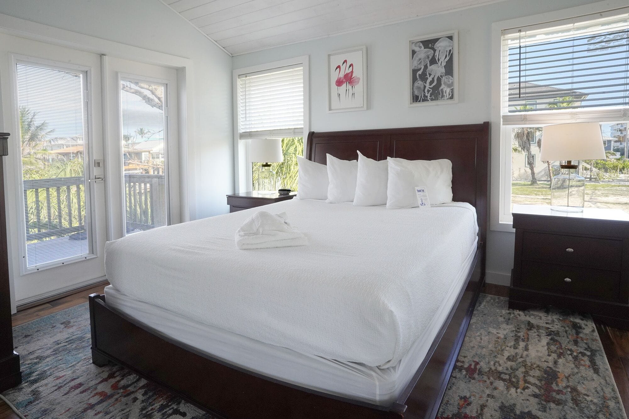 A large bed with white linens, a wooden headboard, and a white towel on the bed. There are two bedside tables with lamps, a window with blinds, and a framed picture on the wall.