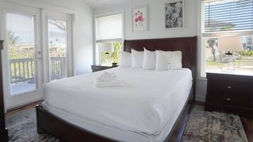 Un grand lit avec du linge de lit blanc, une tête de lit en bois et une serviette blanche posée sur le lit. Il y a deux tables de chevet avec des lampes, une fenêtre avec des stores et un tableau encadré au mur.