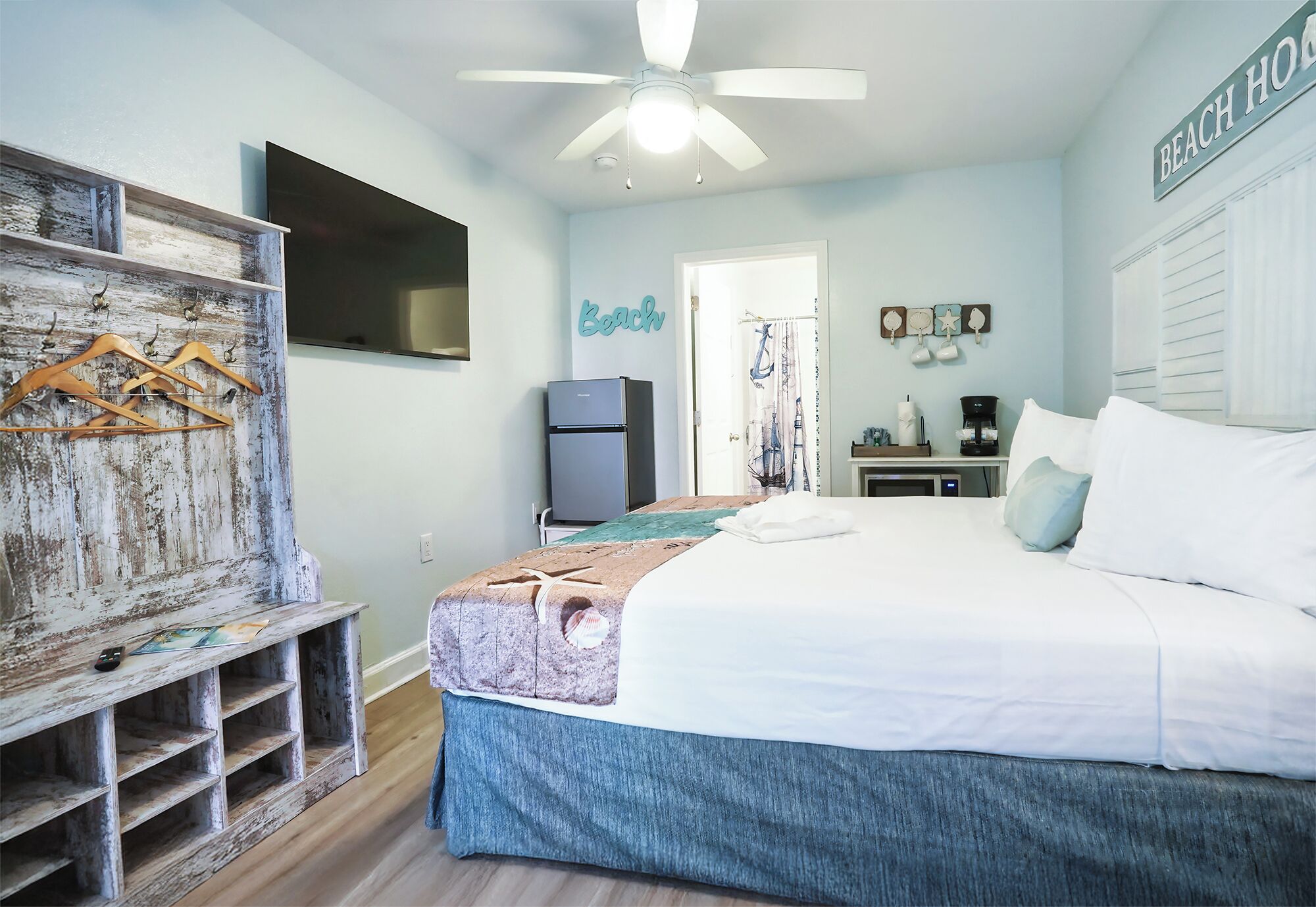 Un dormitorio con cama, ventilador de techo, televisión, refrigerador y un letrero en la pared que dice 'Beach House'.