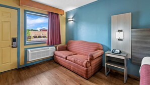 A hotel room with a red sofa, a small bedside table with a phone, a window with curtains, a radiator, and a door. - Econo Lodge Charlotte Airport Area (Charlotte)
