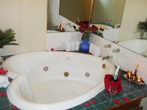 A bathroom with a large bathtub, a mirror, and a sink. There are rose petals scattered on the floor, a blue vase with flowers, and a bottle of champagne on the sink.