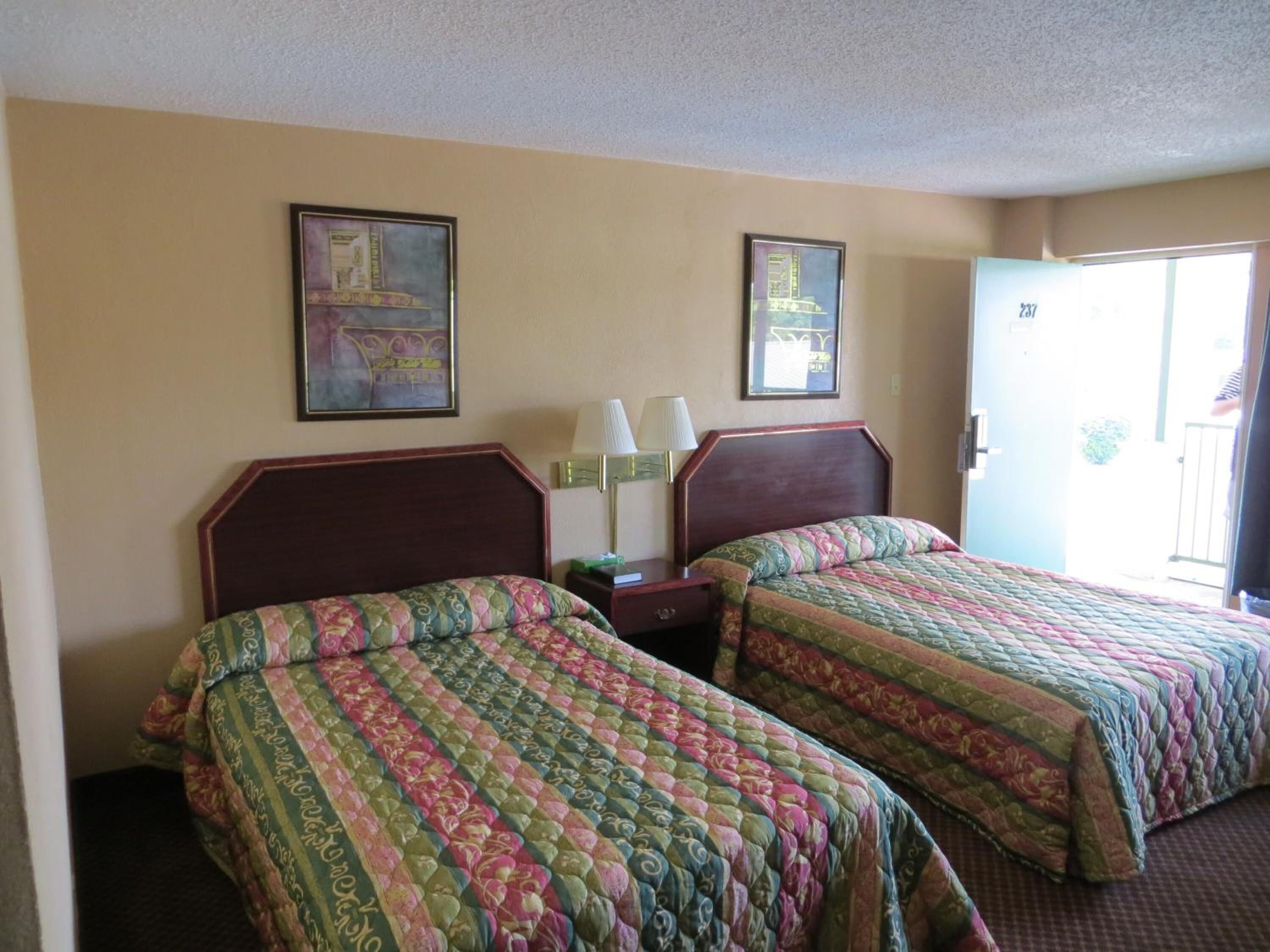 A hotel room with two beds, a nightstand, a lamp, and two framed pictures on the wall.