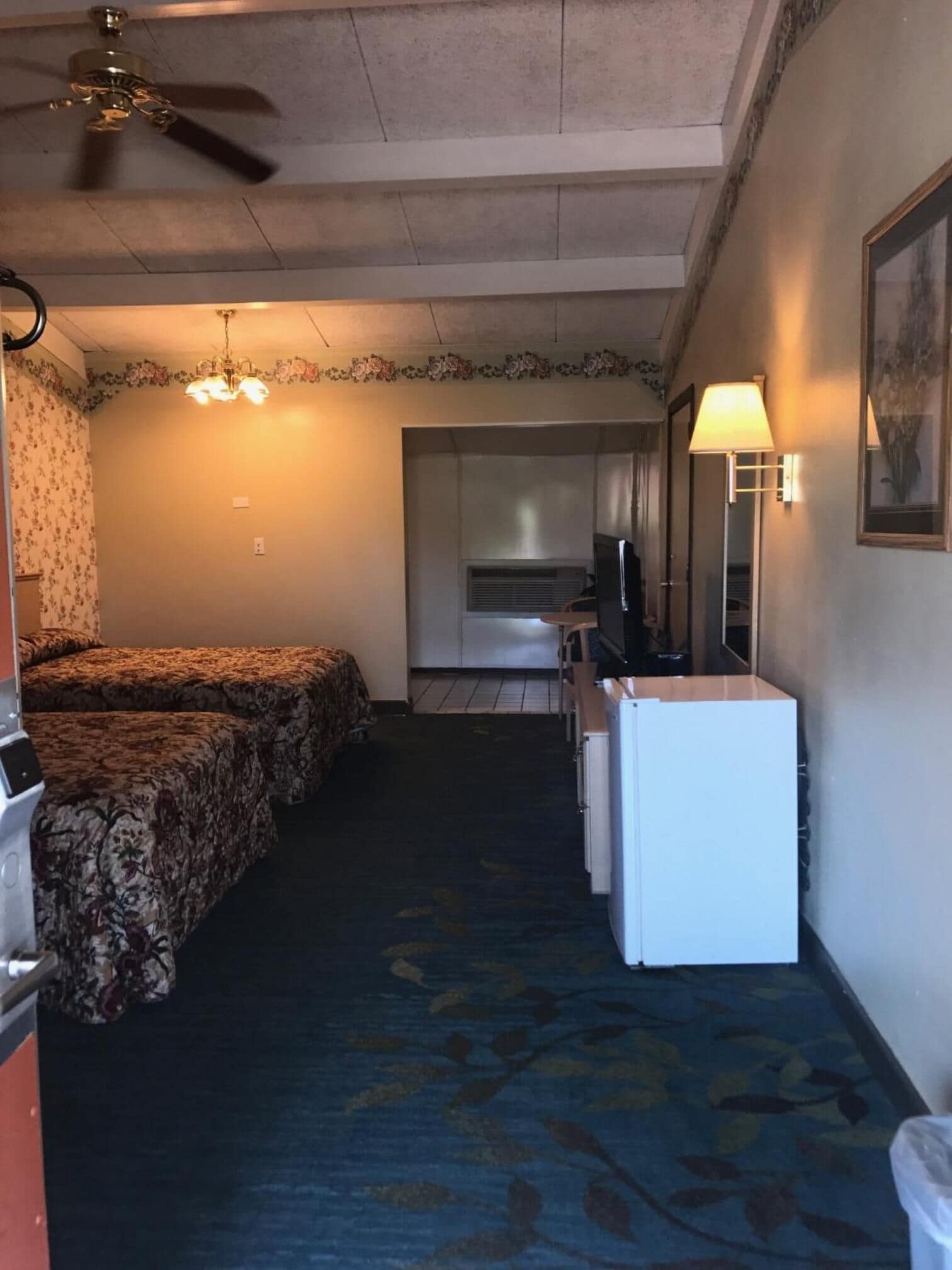 Habitación de hotel con dos camas, ventilador de techo, kitchenette y refrigerador pequeño.