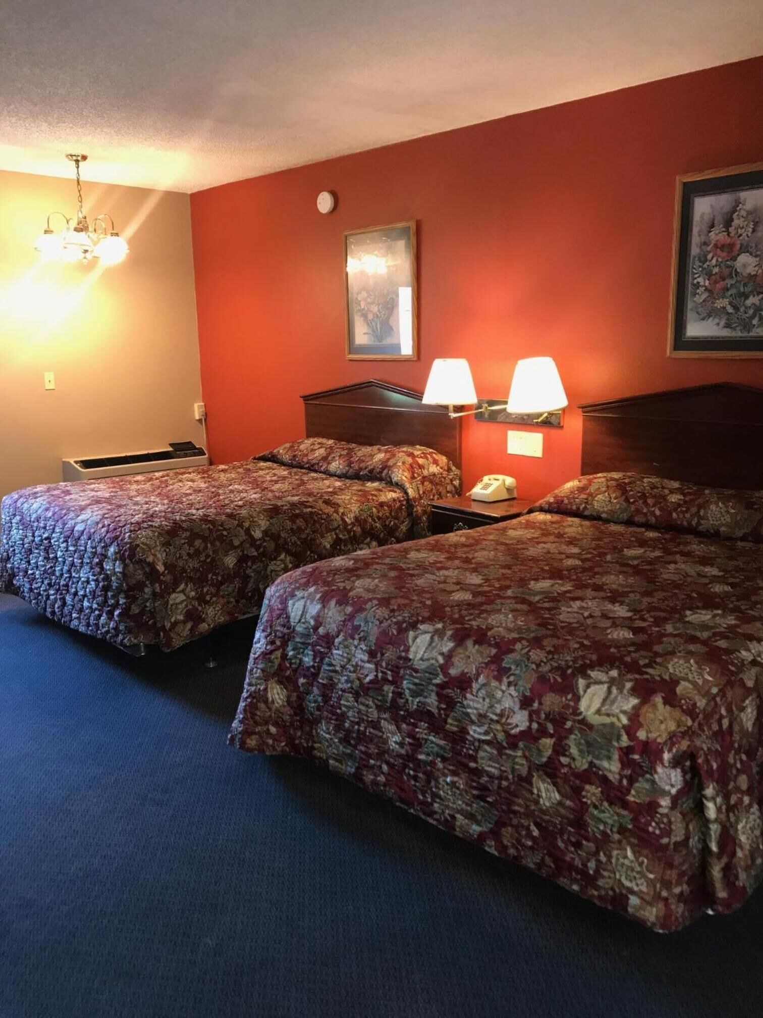 A hotel room with two beds, a nightstand, a lamp, a clock, and a painting on the wall.