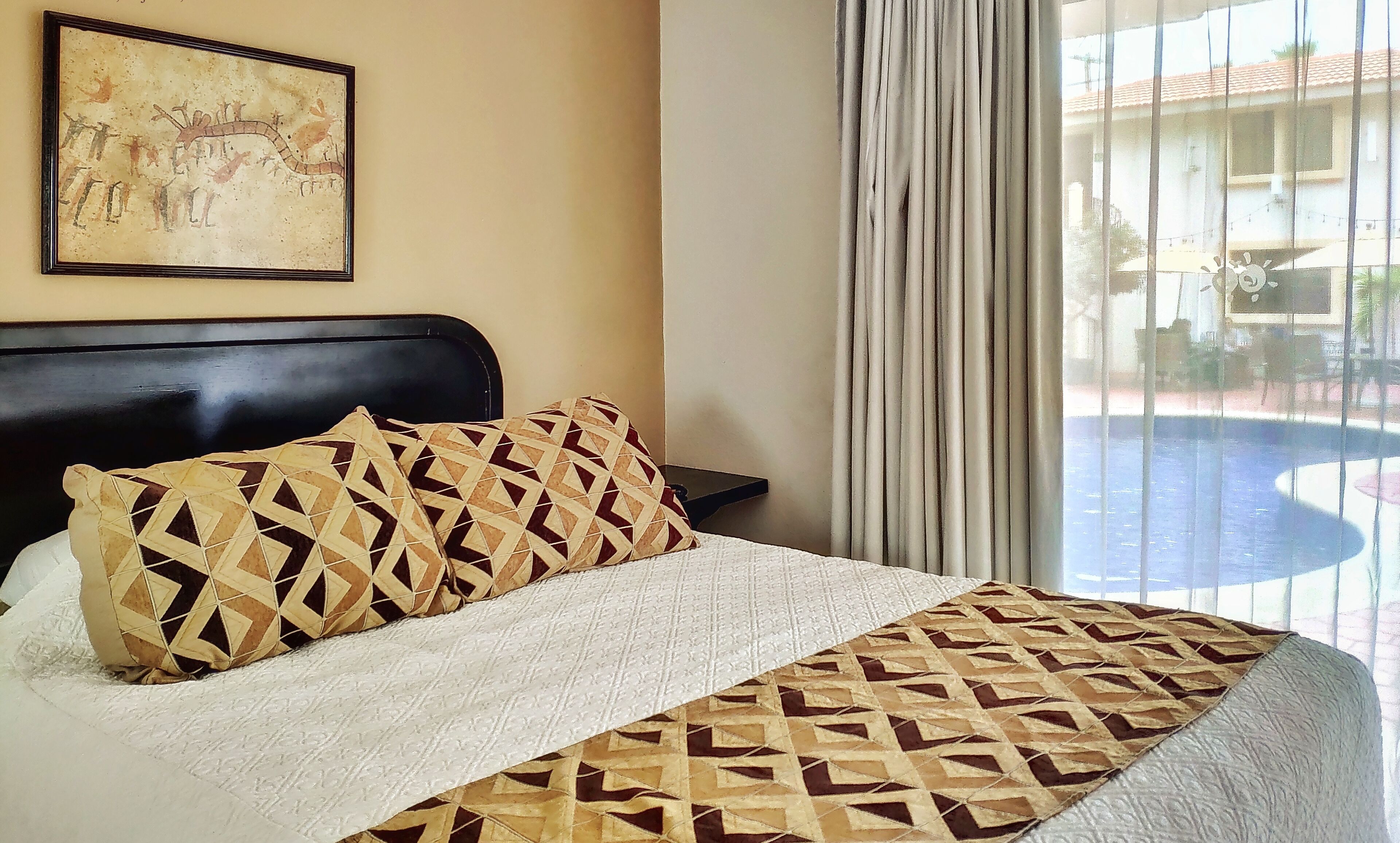 Een hotelkamer met een bed, een schilderij aan de muur en uitzicht op een zwembad.