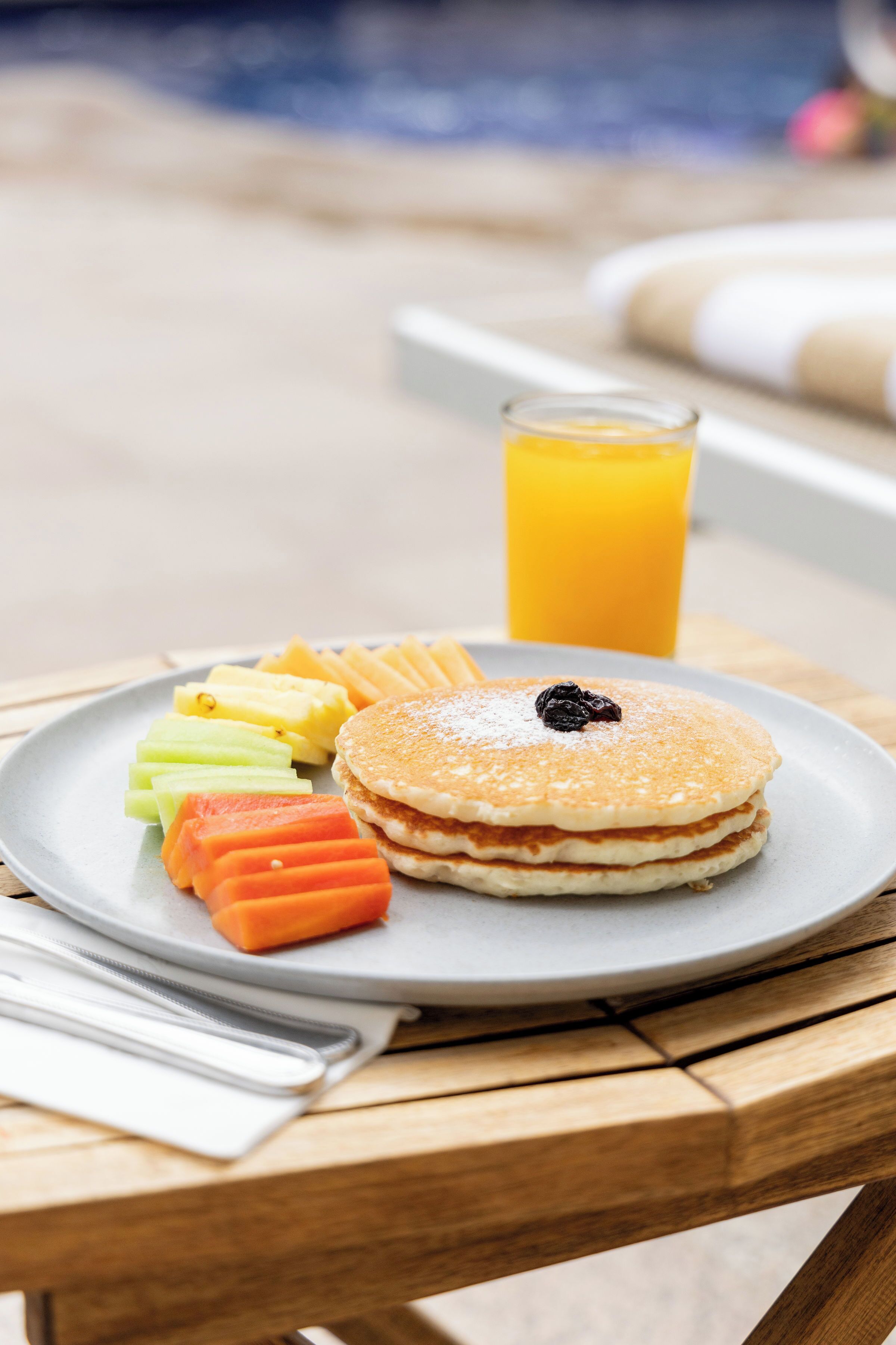 Une assiette de petit-déjeuner avec des crêpes, des fruits et un verre de jus d’orange, posée sur une table en bois.