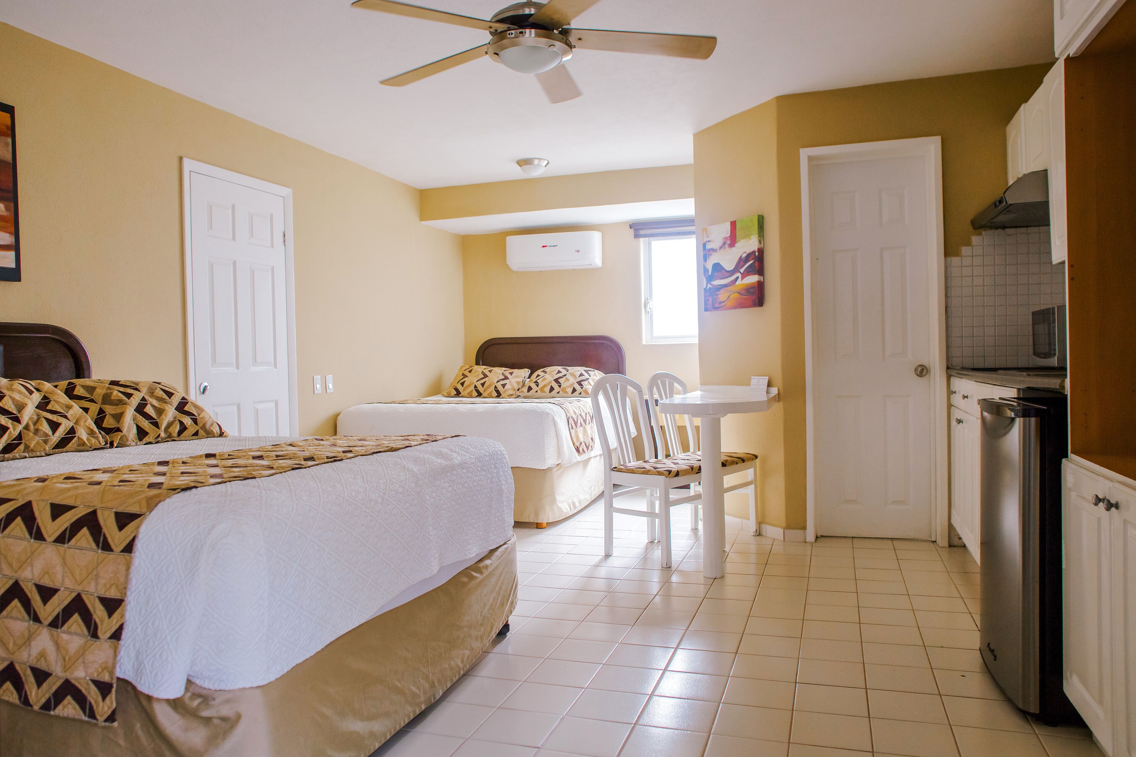 A hotel room with a bed, a desk, a chair, a ceiling fan, and a kitchen area with a microwave.
