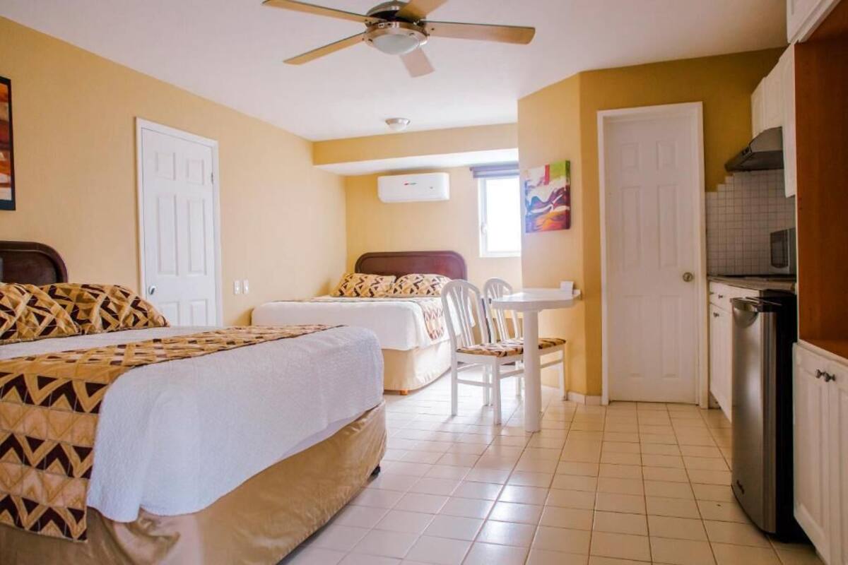 A hotel room with a bed, a desk, a chair, a ceiling fan, and a kitchen area with a refrigerator.