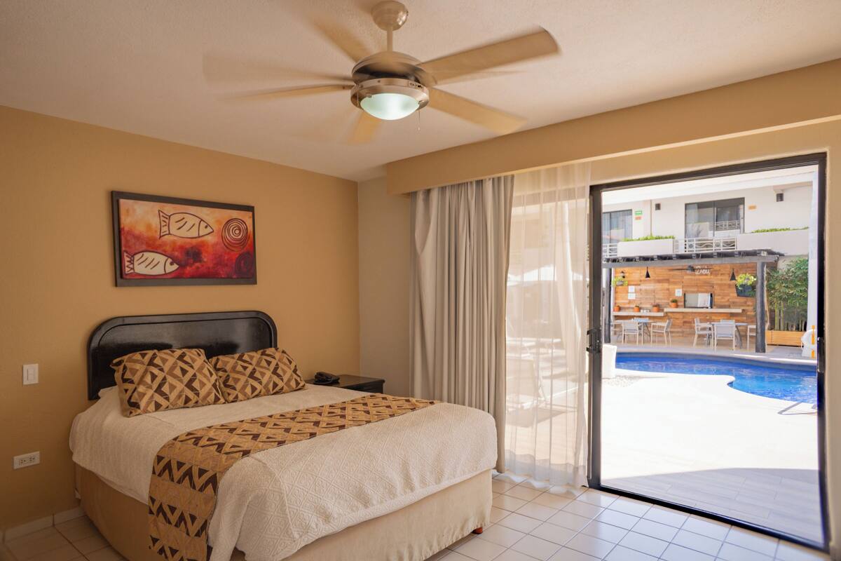 A hotel room with a bed, a ceiling fan, a painting, a sliding glass door to a pool area, and a view of the pool.