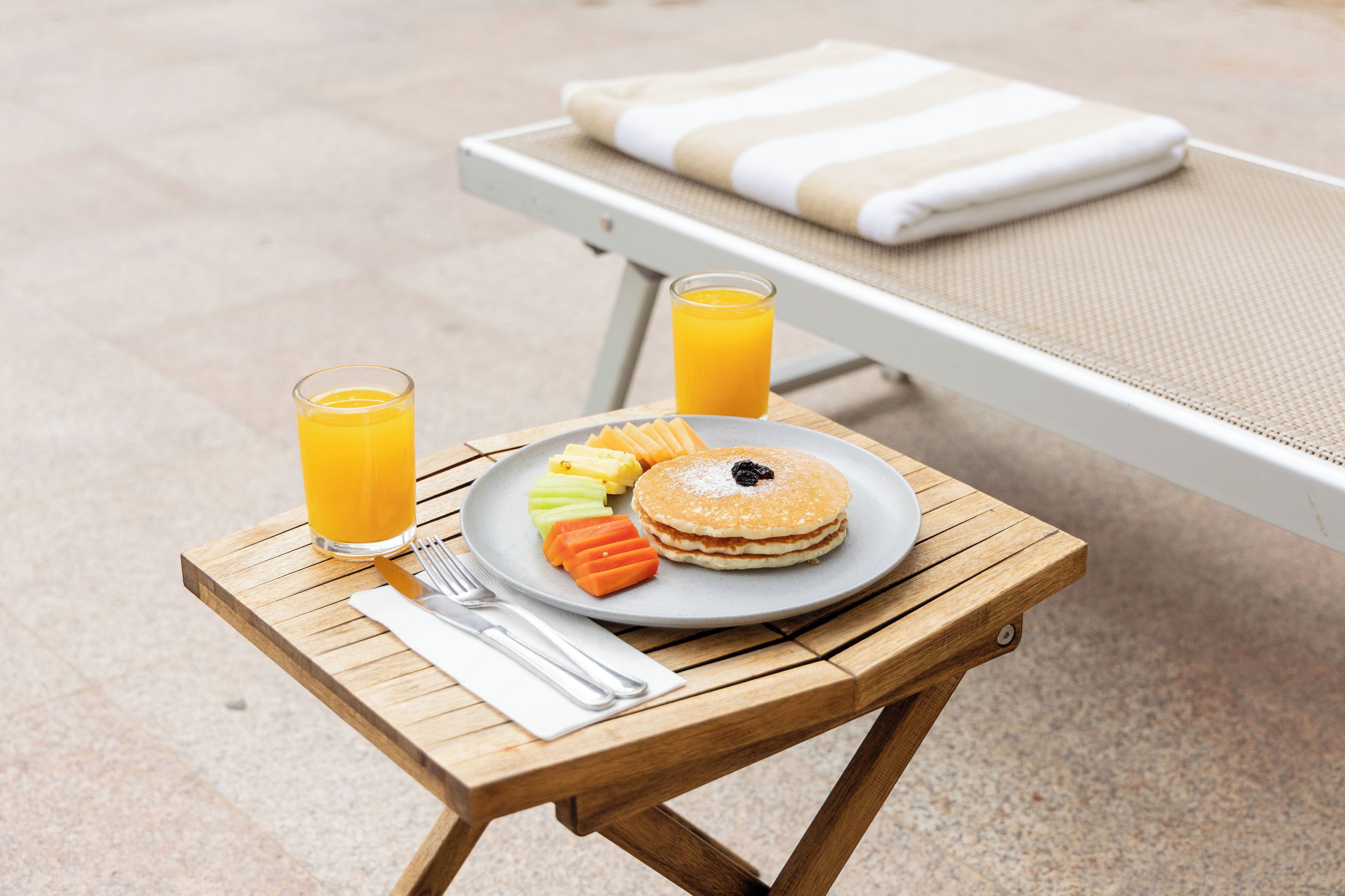 A wooden table with a plate of pancakes, fruit, and a glass of orange juice, set on a beach with a lounge chair in the background.