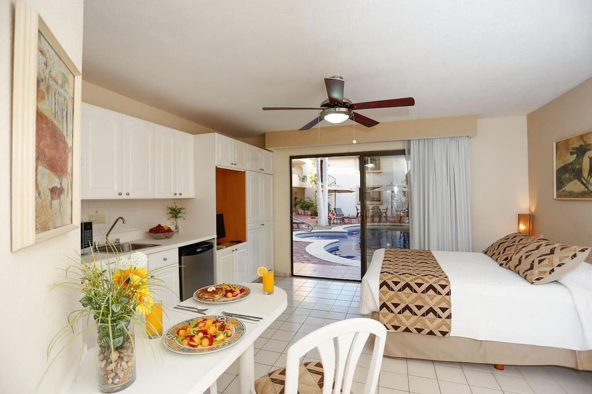 A hotel room with a bed, a desk with breakfast, a ceiling fan, and a view of a pool.