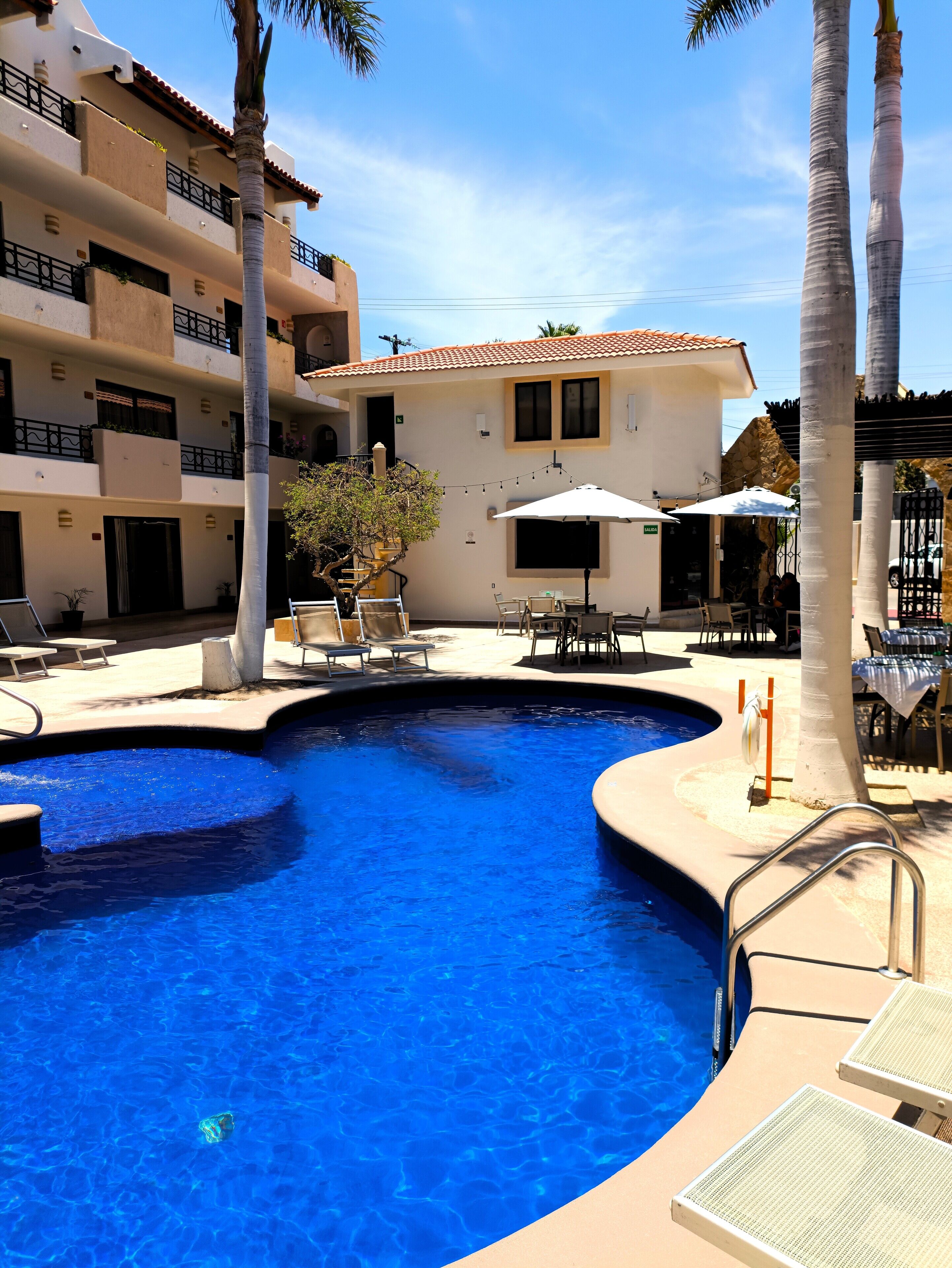 A swimming pool with lounge chairs and umbrellas, surrounded by a building with balconies and a tiled roof.