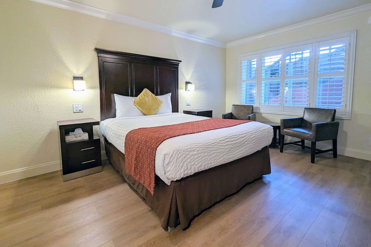 Quarto de hotel com uma cama grande, uma mesa de cabeceira, uma escrivaninha com cadeira e uma janela com persianas.