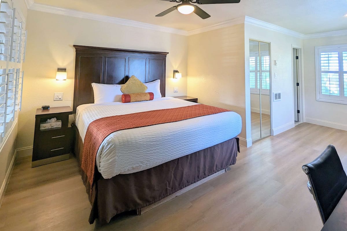 Quarto com uma cama grande, cabeceira de madeira, mesas de cabeceira com luminárias, ventilador de teto e uma porta de vidro deslizante que dá para uma varanda.