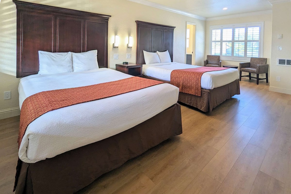 Quarto de hotel com duas camas, cabeceiras de madeira, uma janela com persianas, uma cadeira e uma pequena mesa.