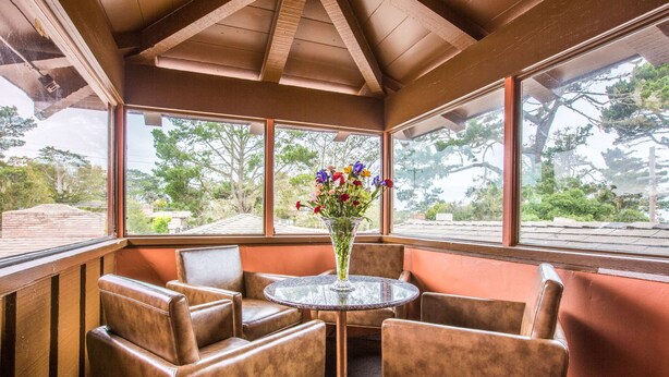 A corner seating area with leather chairs and a glass table, a vase of flowers, and large windows offering a view of trees and buildings. - Clarion Collection Wilkie's Inn Pacific Grove - Monterey (Pacific Grove)