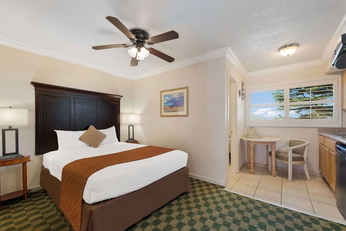 Quarto de hotel com cama, escrivaninha, cadeira, ventoinha no teto, janela com persianas e um quadro na parede.