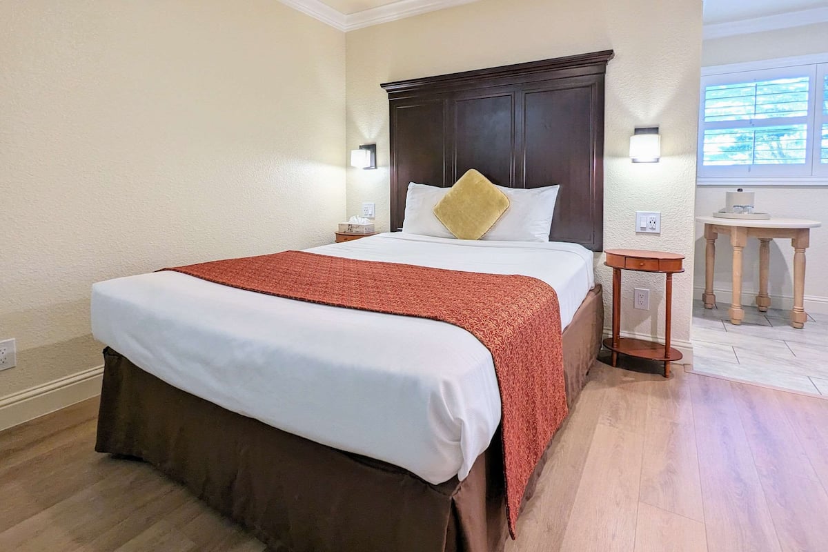Quarto de hotel com uma cama grande, uma mesa de cabeceira de madeira, uma pequena mesa e uma janela com persianas.