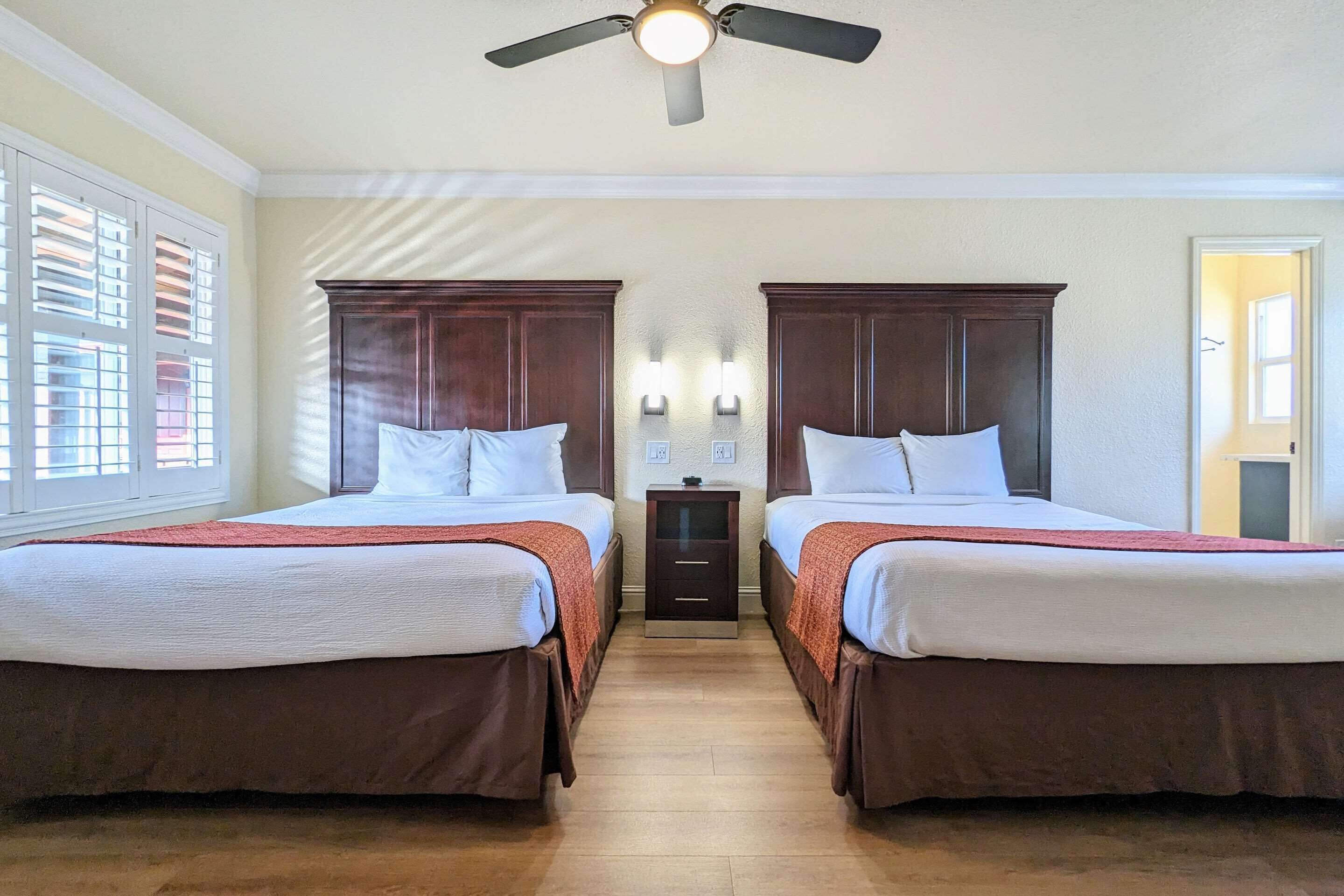 Habitación de hotel con dos camas, un ventilador de techo, una mesita de noche y una ventana con persianas.