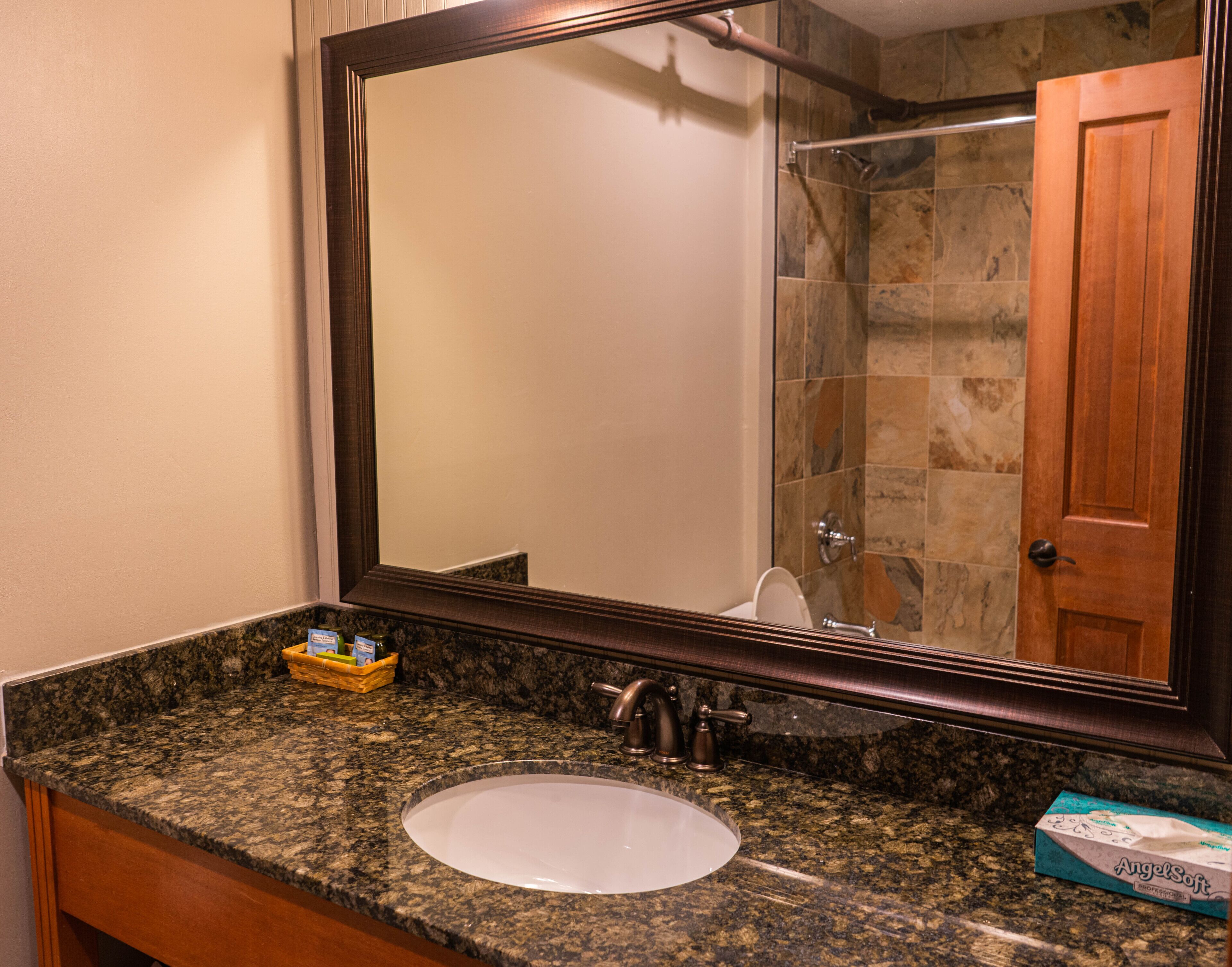 Un baño con encimera de granito, un lavamanos blanco, un espejo grande y una zona de ducha.