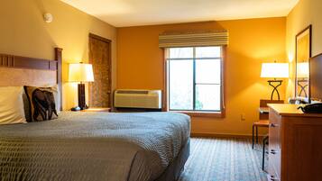 Quarto de hotel com uma cama grande, duas mesas de cabeceira com abajures, um aparador de madeira, uma janela com persianas e um aparelho de ar condicionado.