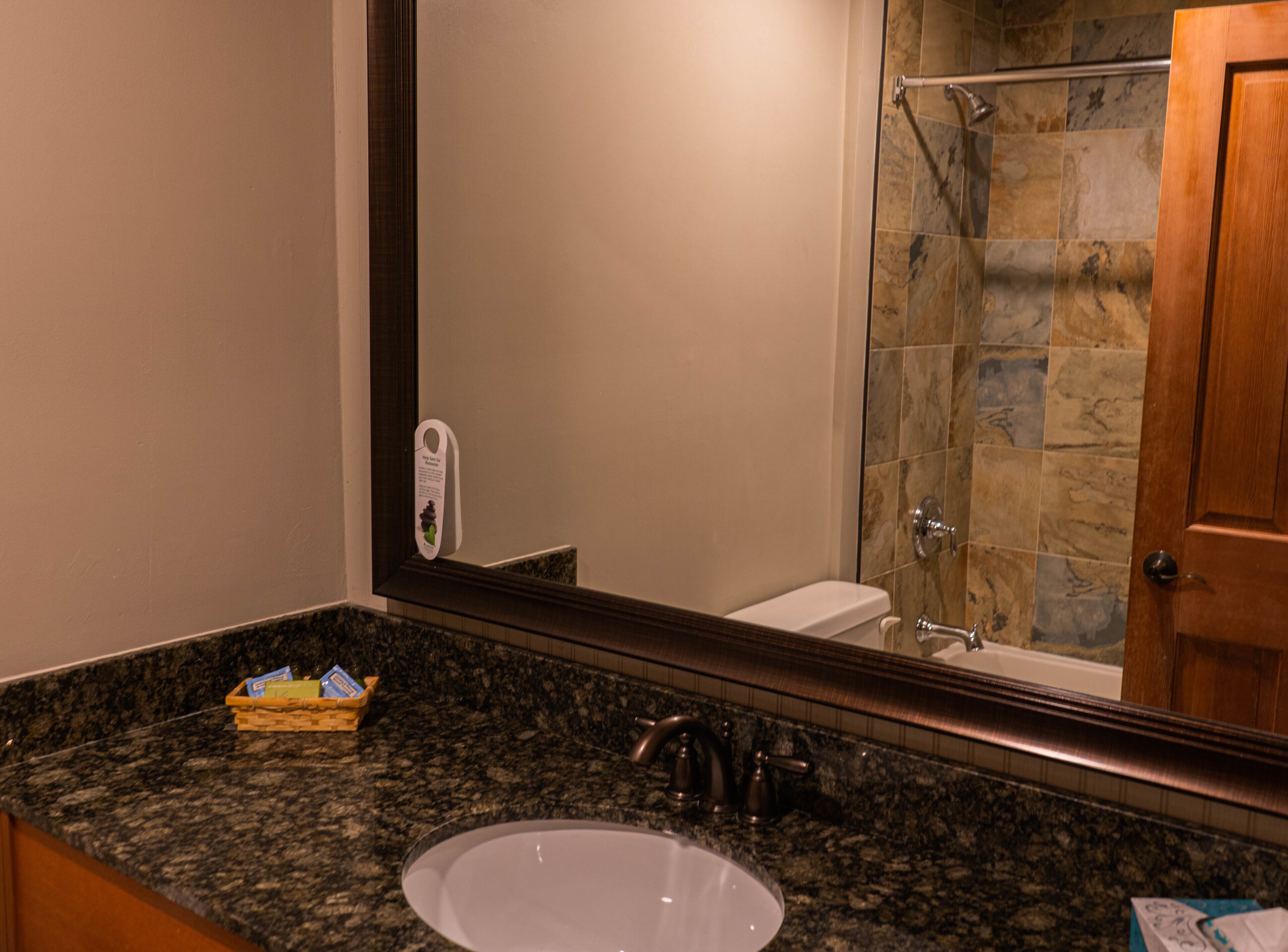Une salle de bain dotée d’un comptoir en granit, d’un lavabo, d’un miroir, d’une douche aux carreaux de pierre et d’une porte en bois.