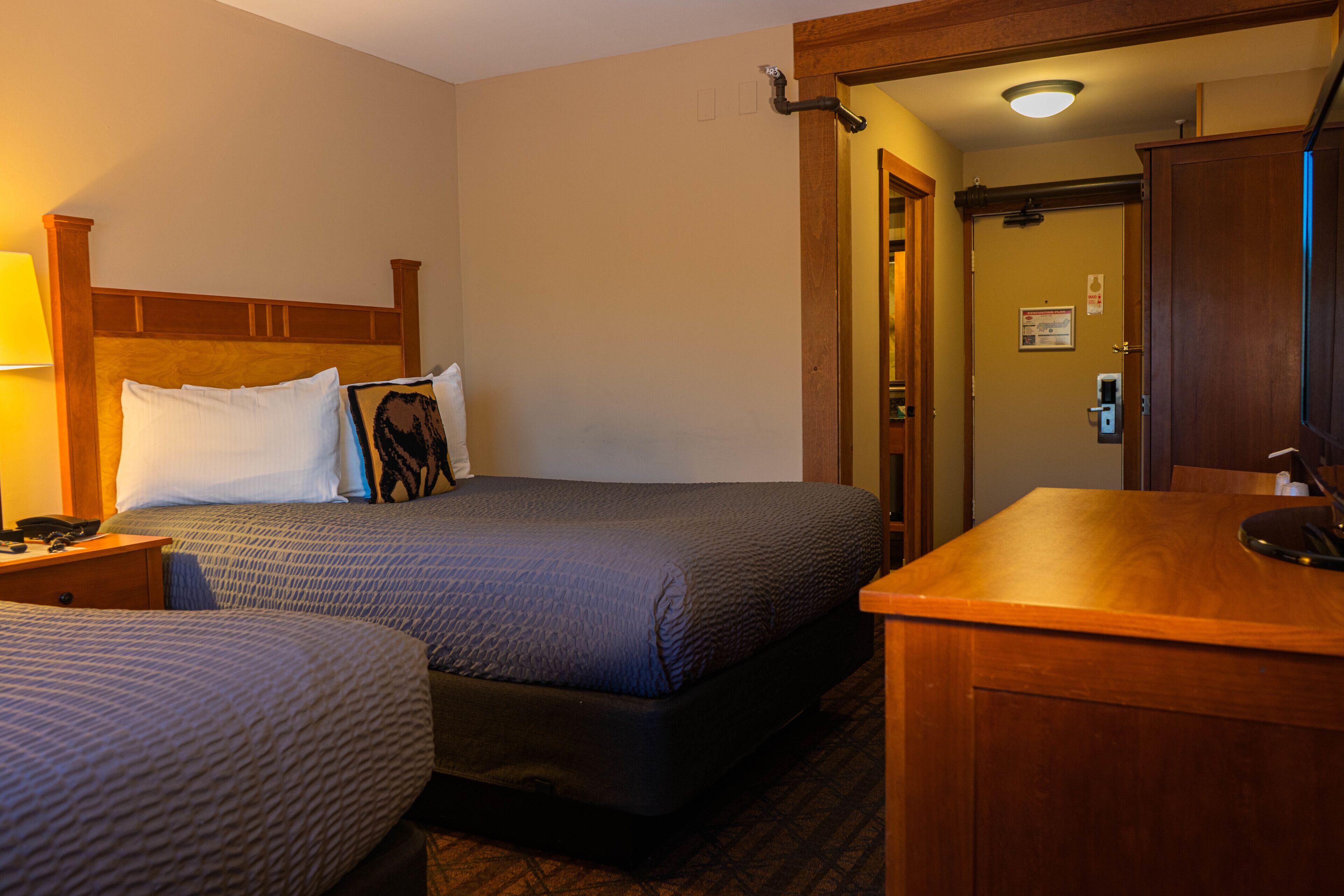 Ein Hotelzimmer mit zwei Betten, einem hölzernen Kopfteil, einem Nachttisch mit Lampe sowie einem Schreibtisch mit Spiegel.
