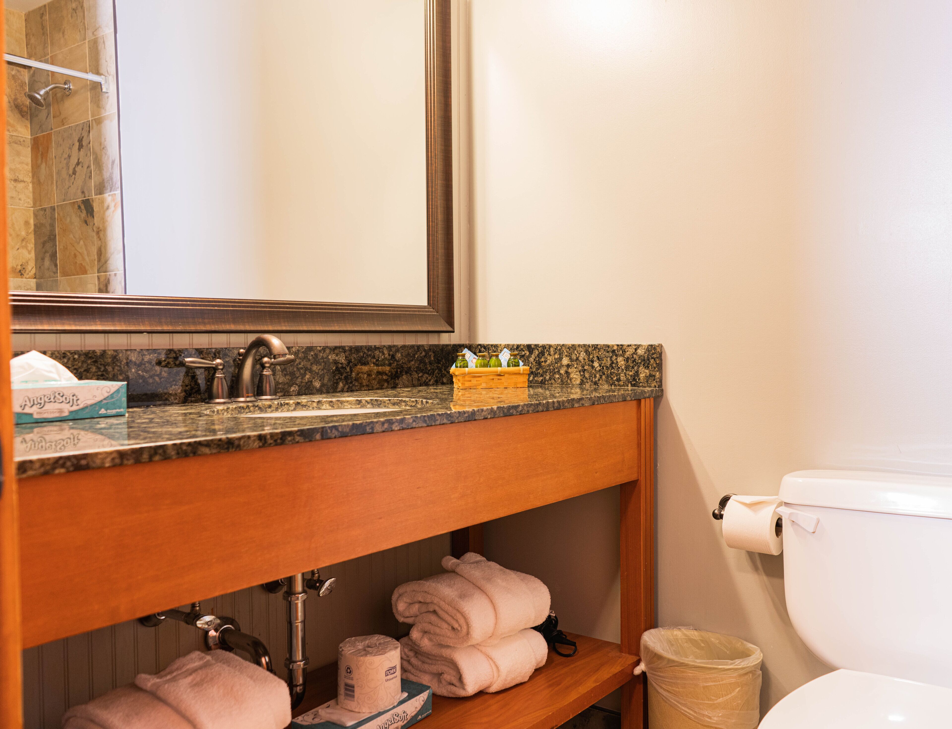 A bathroom with a granite countertop, a mirror, a toilet, and a shelf with towels and toiletries.