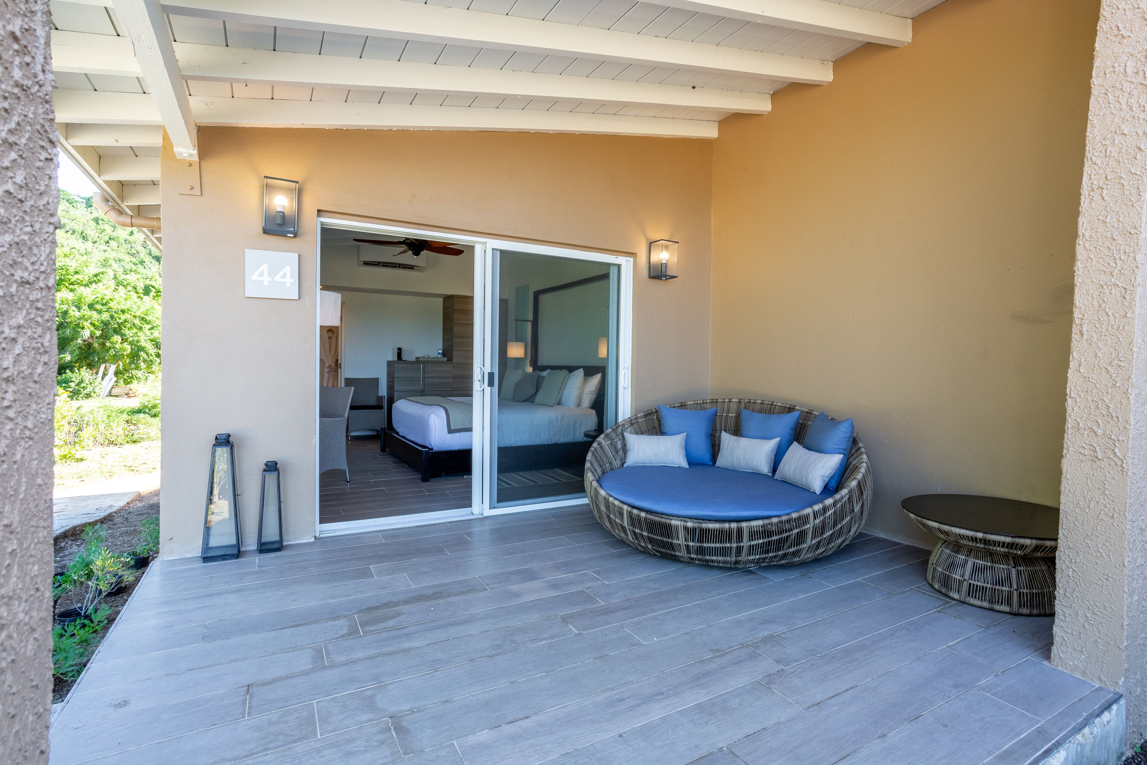A covered patio area with a wicker sofa and a small table, a sliding glass door leading to a bedroom, and a number '44' sign on the wall.