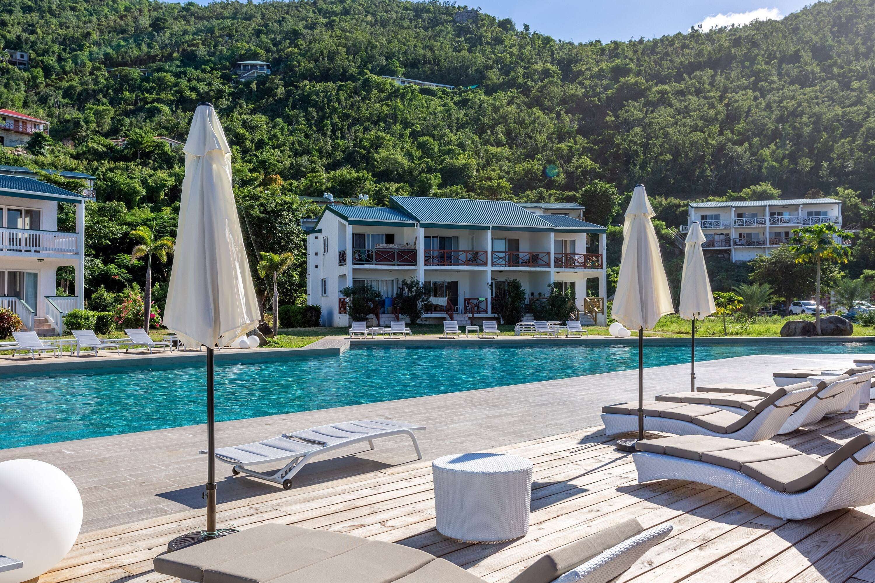 Zona de piscina con sillas de descanso y sombrillas, una terraza de madera y un edificio con balcones al fondo.