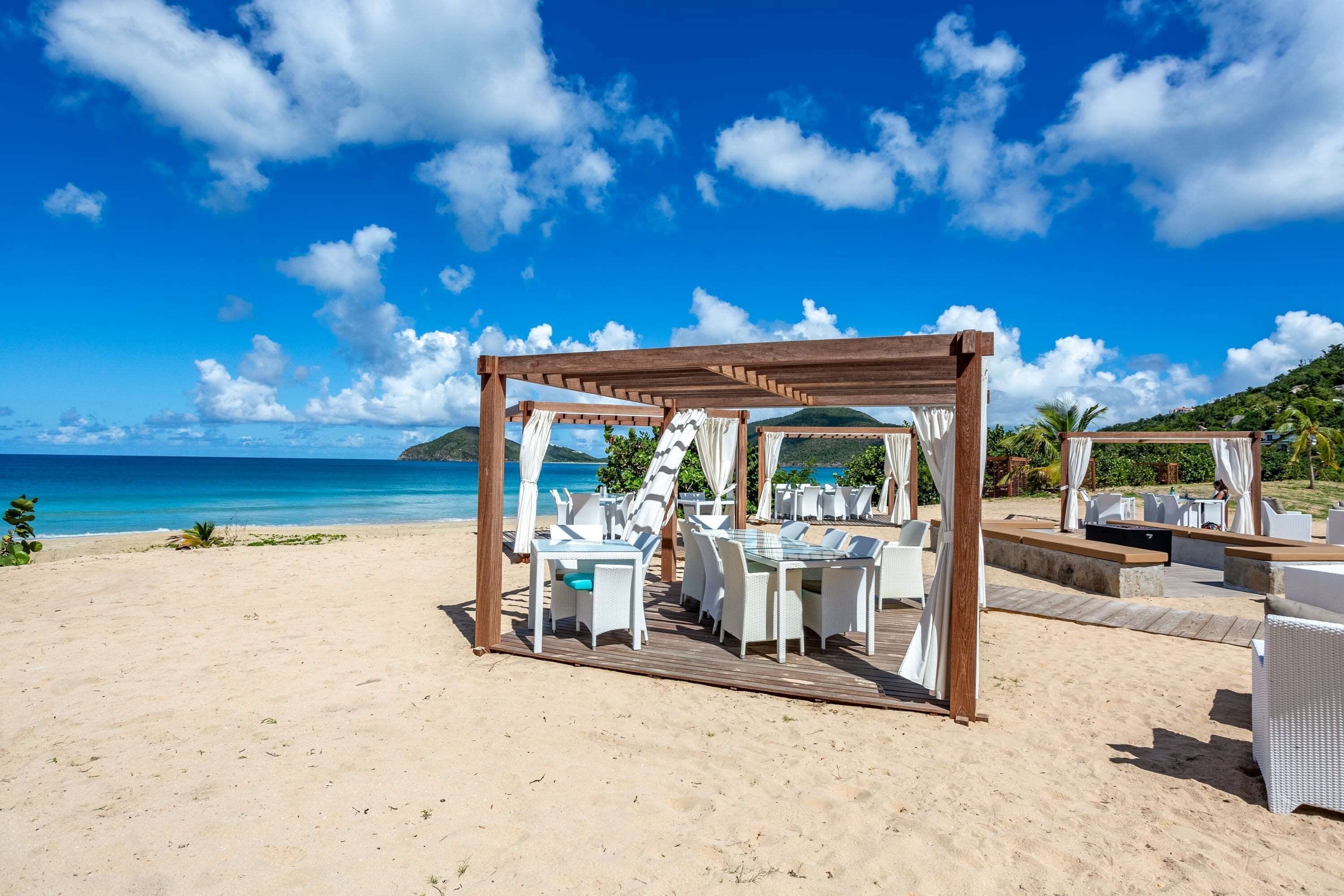 Un espace de restauration en plein air situé au bord de la plage, avec des tables et des chaises, une pergola en bois et une vue sur l’océan.