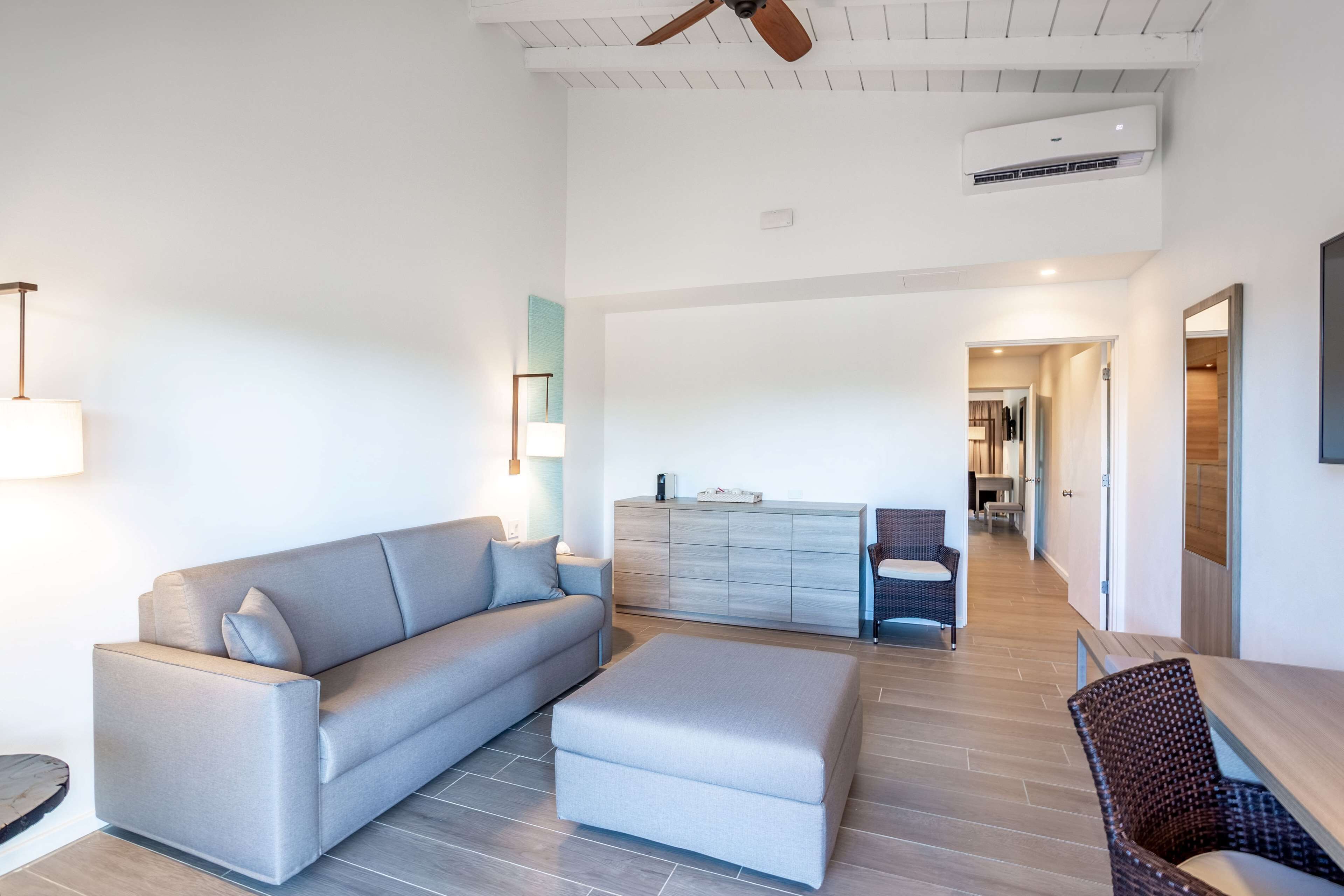 A modern living room with a grey sofa, a matching ottoman, and a dining area with a wooden table and wicker chairs.