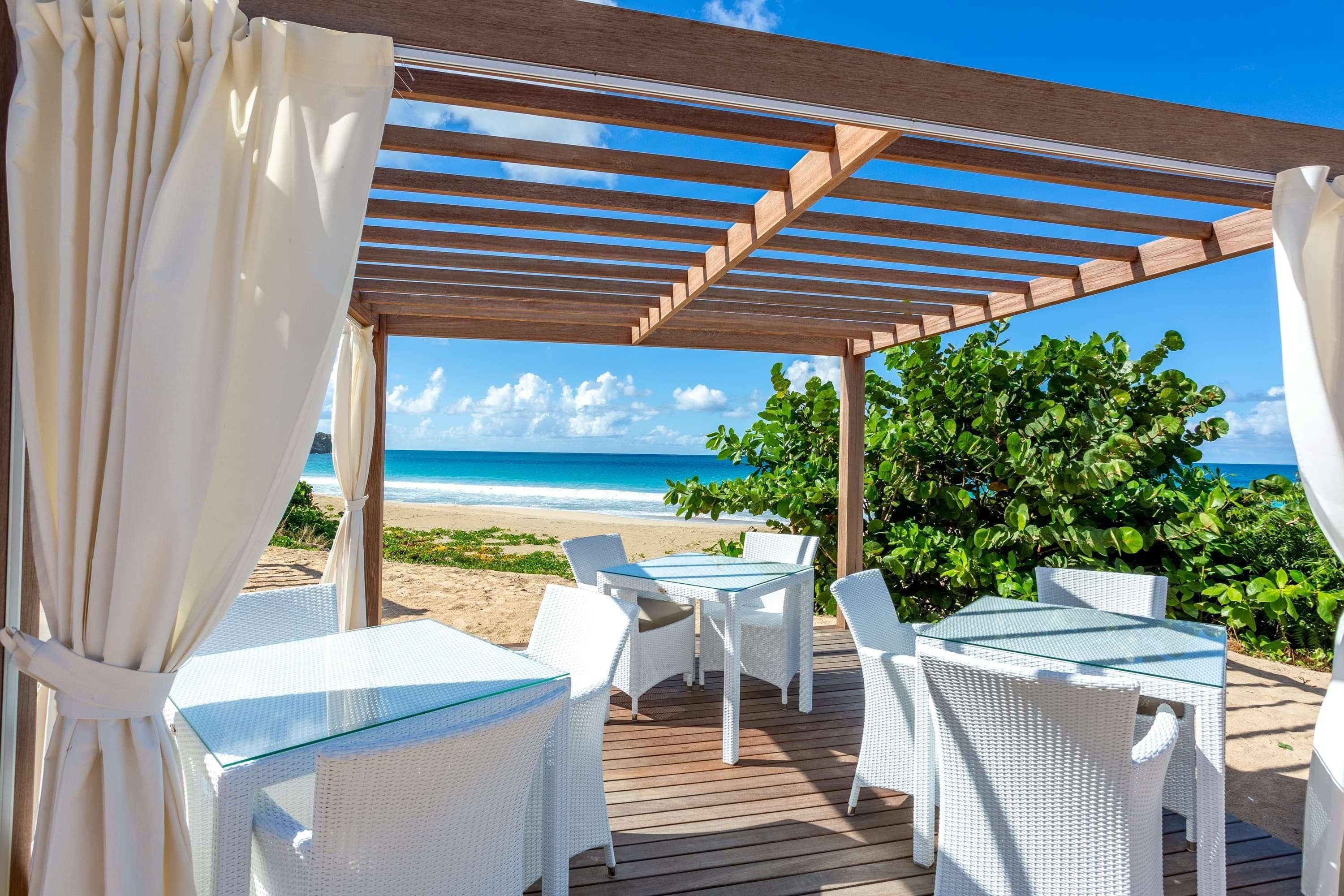 Un patio al aire libre con mobiliario de mimbre blanco y mesas de vidrio, con vista a una playa y al océano.
