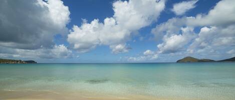 A beach with clear turquoise water, a sandy shore, and a distant island under a partly cloudy sky.