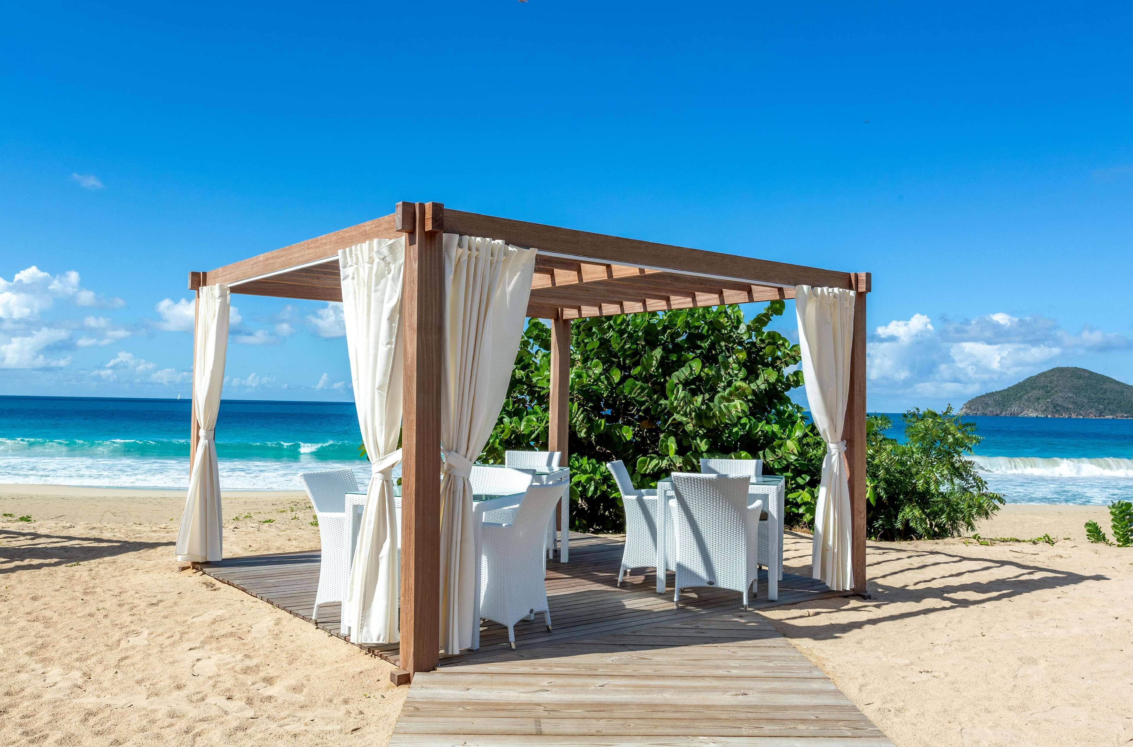 Una pergola in legno con tende bianche e tavolini in vimini bianco su un pontile di legno fronte spiaggia.