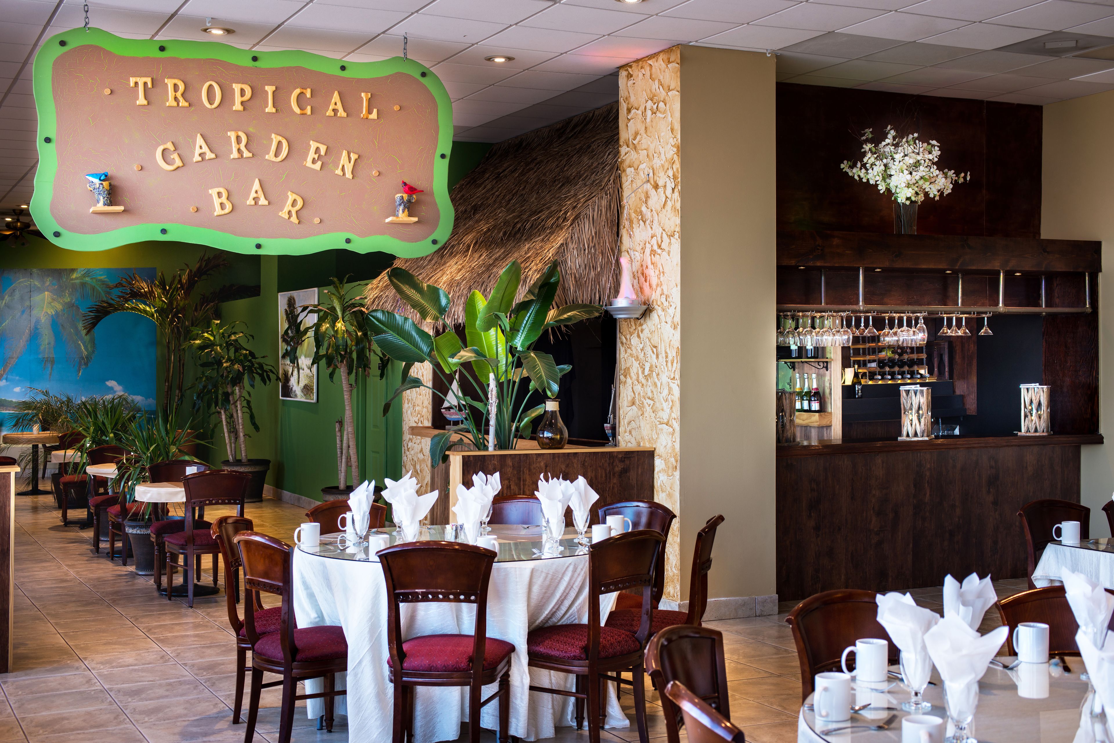 A restaurant with a tropical theme, including a sign reading "Tropical Garden Bar," wooden tables and chairs, and a bar area with bottles and glasses.