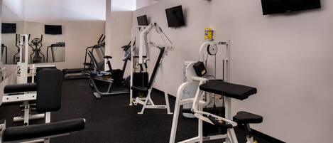 A gym with various exercise machines, a television mounted on the wall, and a mirror reflecting the equipment.