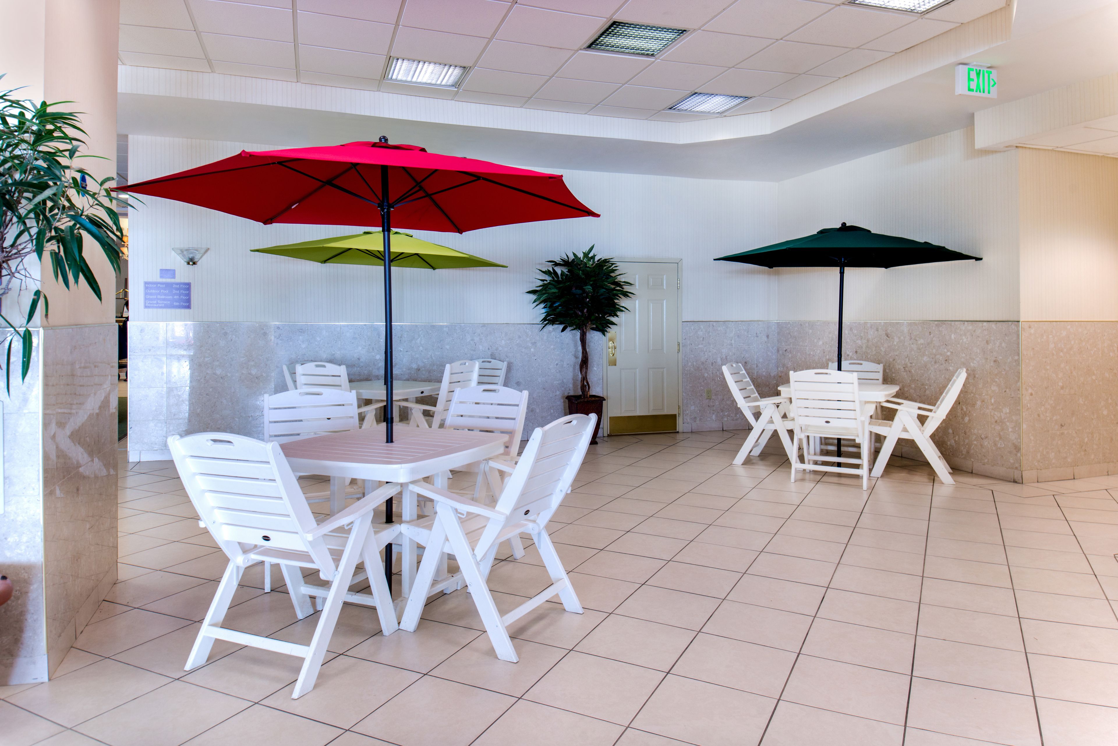 A spacious indoor area with white tables and chairs, red and green umbrellas, and a potted plant.
