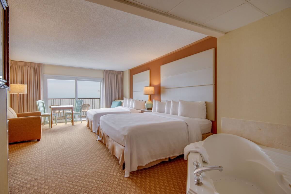 Une chambre d’hôtel avec deux lits, un coin salon comprenant une chaise et une table, un balcon avec des rideaux et une baignoire.