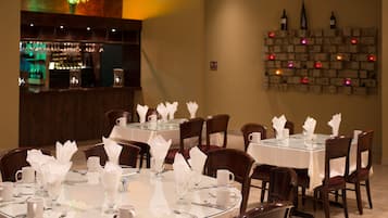 Un restaurant avec des tables rondes disposées pour le service, un bar à l’arrière-plan et un mur décoré de bouteilles et de guirlandes lumineuses.