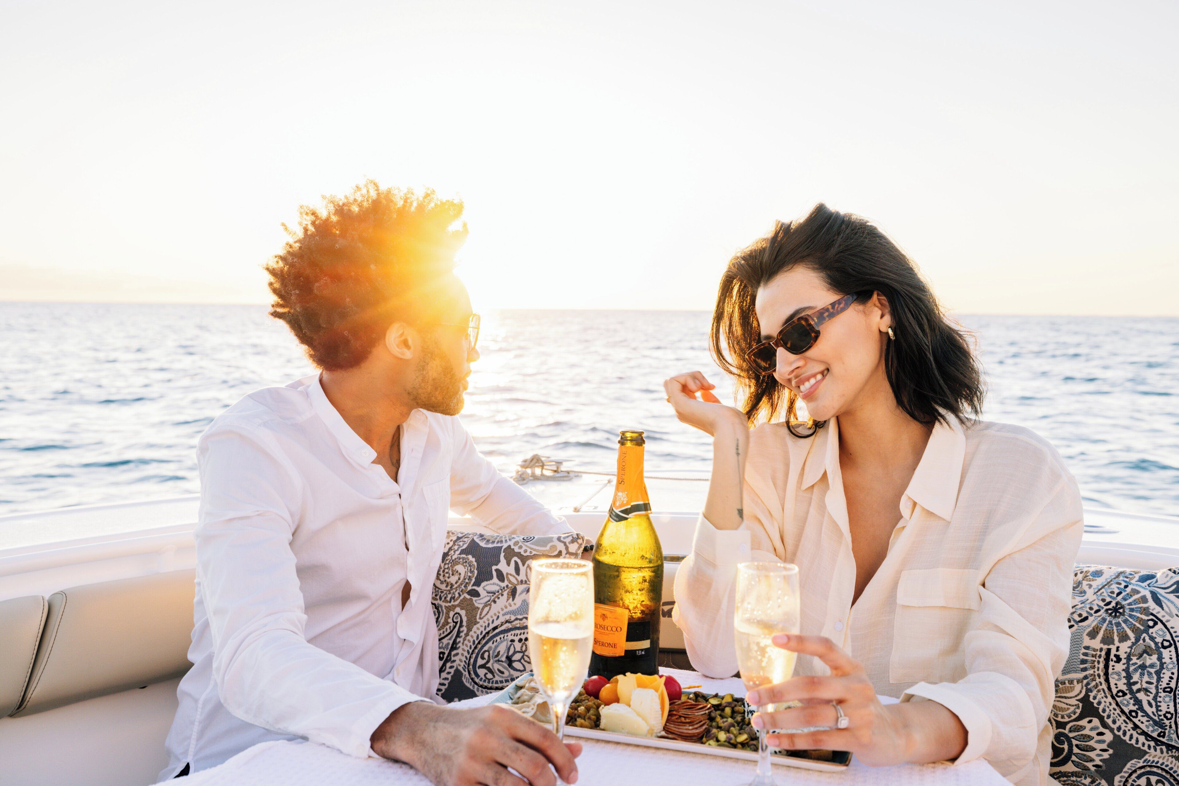 Un couple est à bord d’un bateau, avec une bouteille de champagne et une assiette de collations.