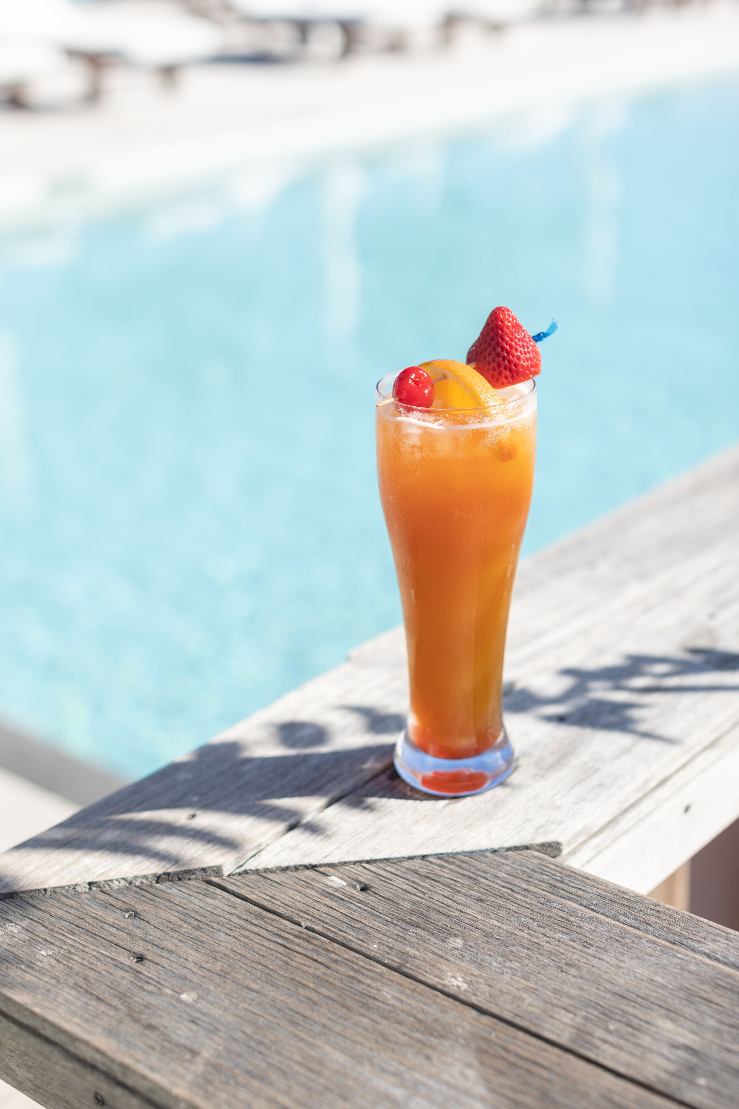 A tall glass with a drink garnished with a strawberry and an orange slice, placed on a wooden deck by a pool.