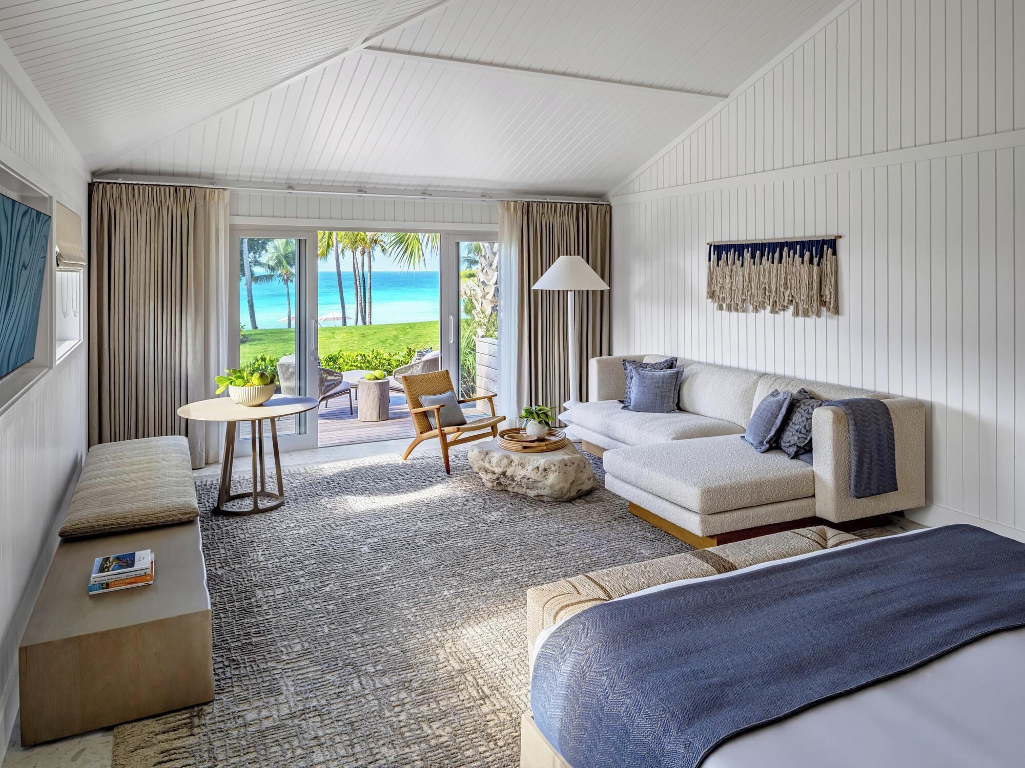 Quarto de hotel com uma janela ampla que oferece vista para a praia, um sofá, uma mesa pequena e uma cadeira.
